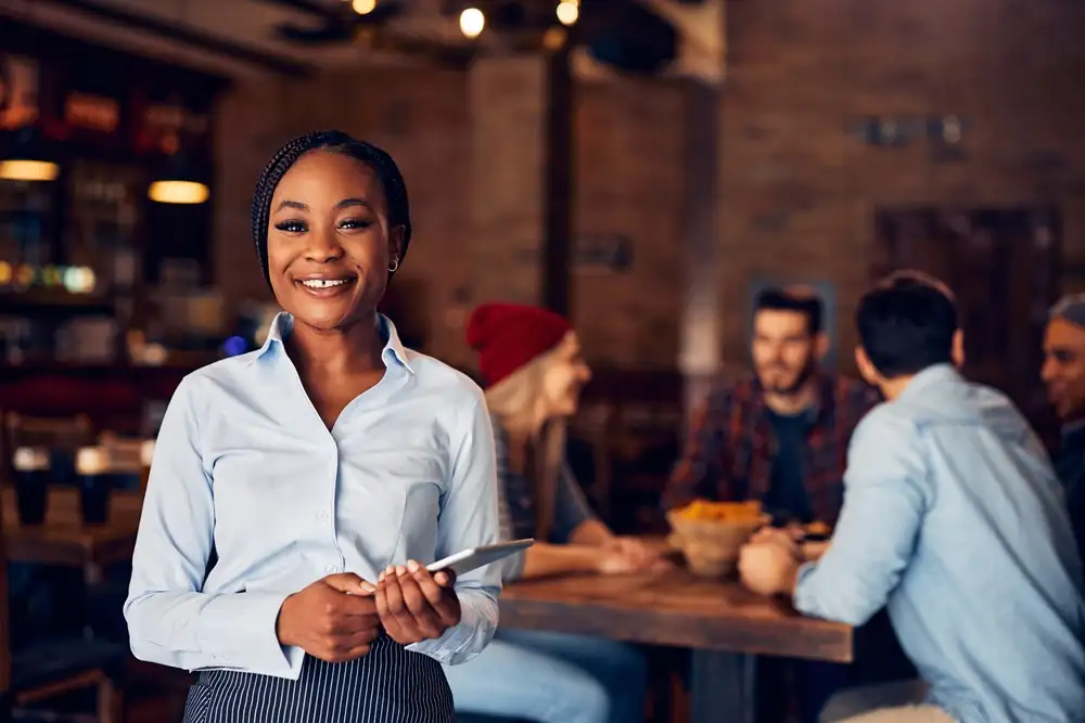 You don't need to be tech-savvy to use restaurant reservation systems. The top systems offer hands-on support, training, and tools to help you succeed, as shown in this image of a restaurant owner smiling as she receives guests at her restaurant.